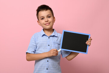 Adorable smiling boy pointing on an empty blank board with a chalk. Space for text on a chalkboard. Pink background with copy space. Concepts of back to school