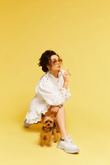 Young glamorous lady in fashionable sunglasses and white dress posing with gold bag and Yorkshire terrier over the yellow background