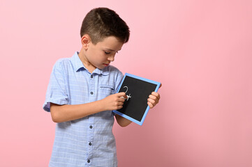 A school boy in blue shirt writes with chalk on a blackboard, isolated over pink background with...