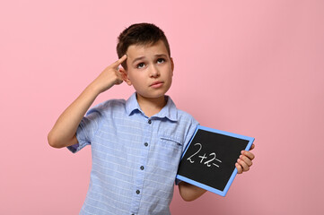 A schoolboy with a blackboard in his hand holds a finger to his temple and with a thoughtful look solves an arithmetic problem, standing on a pink background with a place for text. Back to school.