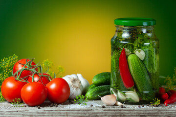 Pickled cucumbers in glass jars and spices and vegetables for preparation of pickles on green background.