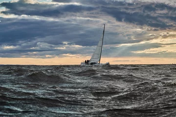 Rolgordijnen The lonely sailboat on the horizon in sea at sunset, the storm sky of different colors, big waves, sail regatta, cloudy weather, only main sail, sun beams © Vladimir Drozdin