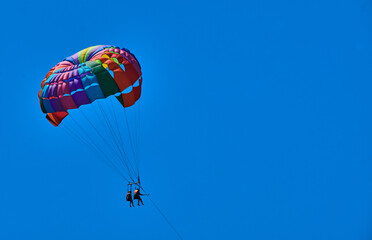 Two people, tourists flying onrainbow-colored parachute in blue sky.