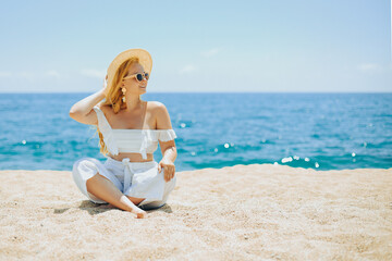 Fototapeta na wymiar Happy girl in white outfit sits on sand against the backdrop of the sea or ocean beach. Woman smiles and laughs, vacation and joy. Fashion model, beautiful jewelry, earrings, hat, sun glasses
