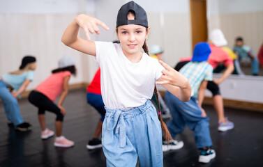 Fototapeta na wymiar Little girl hip hop dancer in casual clothes and cap exercising with friends at dance center