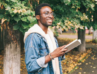 Portrait of young african man student with a book looking away wearing an eyeglasses in autumn city...