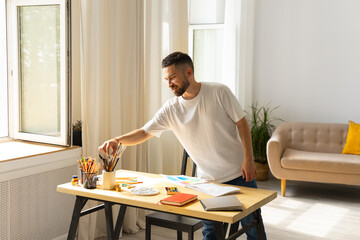 Cuddly caucasian white male artist with dark hair and beard in a white t-shirt takes hands a brush from a plaster glass with Colored pencils on a wooden table in order to start drawing. Modern room