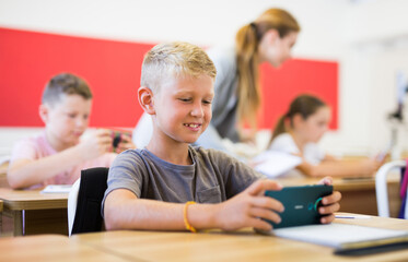 Small school child boy using smartphone while working in classroom during lesson in primary school