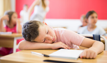 Fototapeta na wymiar Tired preteen schoolboy sleeping at desk in classroom during lesson on blurred background of classmates