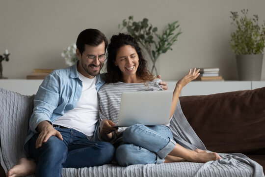 Happy millennial couple using laptop for video call from home, looking at screen, talking at webcam, smiling, laughing, relaxing on couch, speaking with gestures. Family distance communication