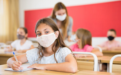 Focused preteen schoolgirl in protective face mask studying in classroom. Necessary precautions in...
