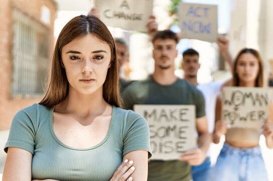 Young hispanic activist woman with arms crossed gesture standing with a group of protesters holding banner protesting at the city.