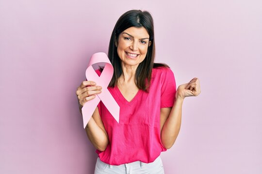 Young hispanic woman holding pink cancer ribbon screaming proud, celebrating victory and success very excited with raised arm