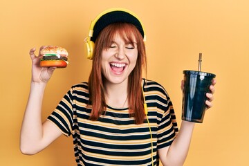 Redhead young woman eating a burger and drinking soda wearing headphones smiling and laughing hard out loud because funny crazy joke. - obrazy, fototapety, plakaty