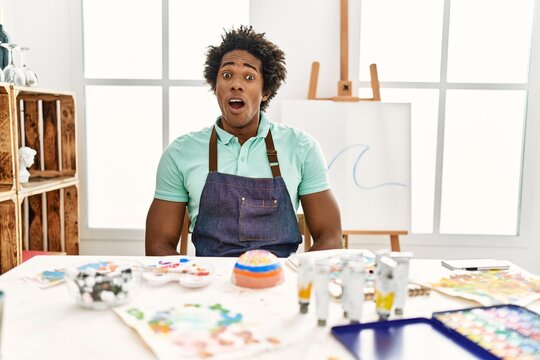 Young african american man sitting on the table at art studio scared and amazed with open mouth for surprise, disbelief face