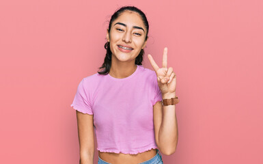 Hispanic teenager girl with dental braces wearing casual clothes smiling looking to the camera showing fingers doing victory sign. number two.