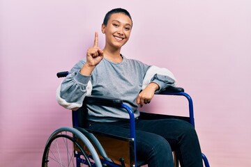 Beautiful hispanic woman with short hair sitting on wheelchair showing and pointing up with finger...