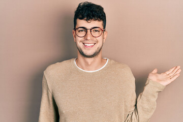 Young hispanic man wearing casual clothes and glasses smiling cheerful presenting and pointing with palm of hand looking at the camera.