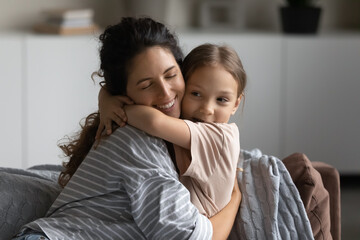 Happy excited mother embracing cute daughter kid with closed eyes. Smiling mom and sweet little girl hugging on couch with love, affection, gratitude. Motherhood, childhood, family