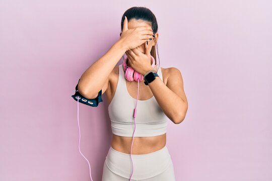 Young hispanic woman wearing gym clothes and using headphones covering eyes and mouth with hands, surprised and shocked. hiding emotion
