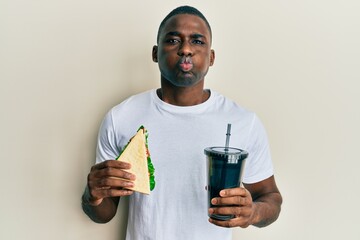Young african american man eating sandwich and drinking soda puffing cheeks with funny face. mouth...