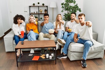 Group of people eating snack sitting on the sofa at home pointing with finger to the camera and to you, confident gesture looking serious