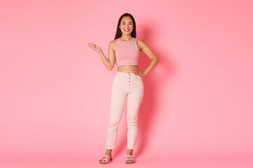 Obraz na płótnie Canvas Fashion, beauty and lifestyle concept. Full-length portrait of attractive tall asian girl smiling and holding left arm raised, demonstrating product over pink background