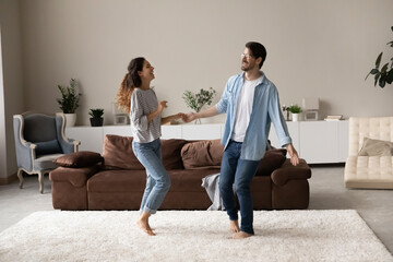 Happy excited millennial couple dancing together in new stylish cozy living room. Husband and wife...