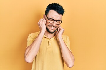 Young hispanic man wearing casual clothes and glasses covering ears with fingers with annoyed expression for the noise of loud music. deaf concept.