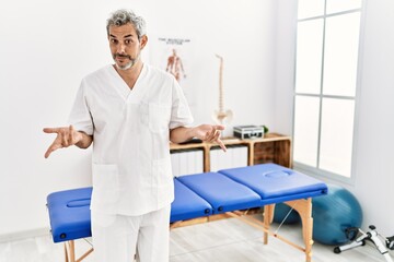 Middle age hispanic therapist man working at pain recovery clinic clueless and confused with open arms, no idea concept.