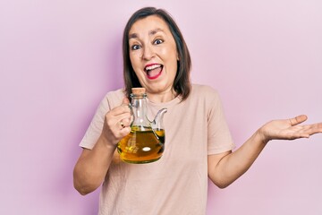 Middle age hispanic woman holding olive oil can celebrating achievement with happy smile and winner expression with raised hand