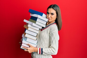 Young brunette student girl holding a pile of books clueless and confused expression. doubt concept.