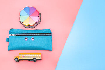 School supplies on pink and blue.
A set of accessories from: school bus, pencil case and paint with rainbow sponges lie on the left on a pink and blue background with space for text on the right,.