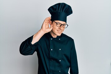 Handsome young man wearing professional cook uniform and hat smiling with hand over ear listening and hearing to rumor or gossip. deafness concept.