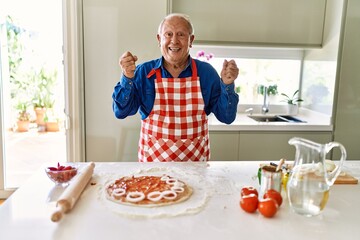 Senior man with grey hair cooking pizza at home kitchen screaming proud, celebrating victory and success very excited with raised arms