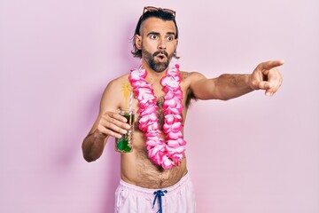 Young hispanic man wearing swimsuit and hawaiian lei drinking tropical cocktail pointing with...