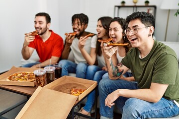 Group of young friends smiling happy eating italian pizza sitting on the sofa at home.