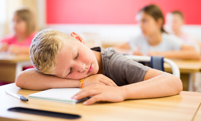 Tired preteen schoolboy sleeping at desk in classroom during lesson on blurred background of classmates