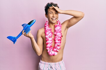 Young handsome man wearing swimsuit and hawaiian lei holding airplane toy stressed and frustrated...