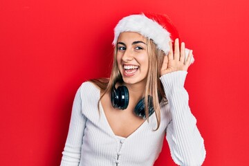 Beautiful hispanic woman wearing christmas hat and headphones smiling with hand over ear listening and hearing to rumor or gossip. deafness concept.