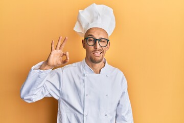 Bald man with beard wearing professional cook apron doing ok gesture winking looking at the camera...