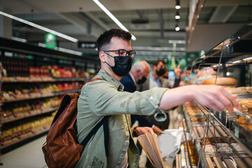 Young man buying freshly baked pastry in big modern bakery. He is wearing face protective mask due to Coronavirus epidemic.
