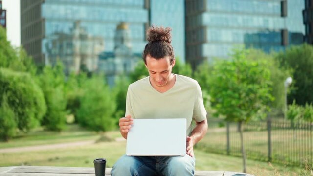 Zoom in slow motion shot of young man working remotely on laptop, closing pc, looking at camera and smiling sitting on bench outdoors. Entrepreneur, freelancer or student finishing work and posing