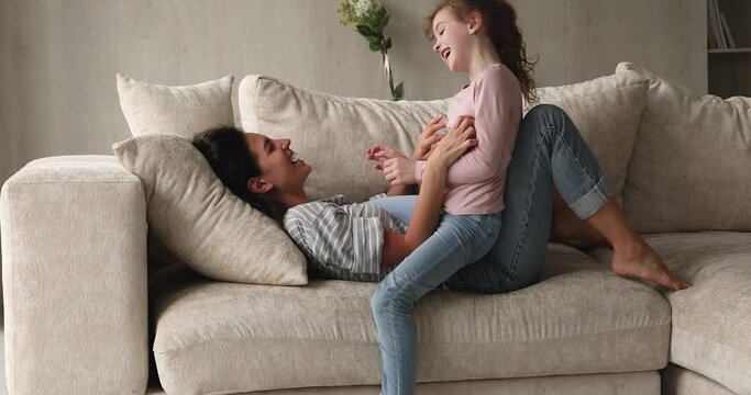 Sincere millennial latino mum or nanny lying on cozy sofa, having fun tickling laughing little preteen kid daughter. Happy different generations family entertaining, enjoying playtime on weekend.