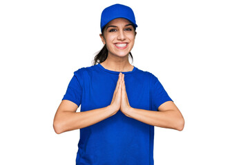 Young hispanic girl wearing delivery courier uniform praying with hands together asking for forgiveness smiling confident.