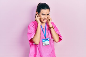 Young brunette woman wearing doctor uniform and stethoscope covering ears with fingers with annoyed expression for the noise of loud music. deaf concept.