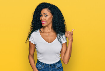 Middle age african american woman wearing casual white t shirt smiling with happy face looking and pointing to the side with thumb up.
