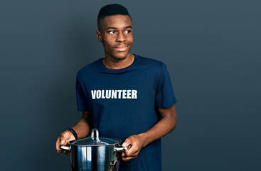Young african american man wearing volunteer holding cooking pot smiling looking to the side and...