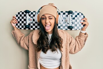 Young hispanic woman holding skate wearing headphones sticking tongue out happy with funny...