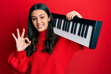 Young hispanic woman holding piano keyboard doing ok sign with fingers, smiling friendly gesturing...